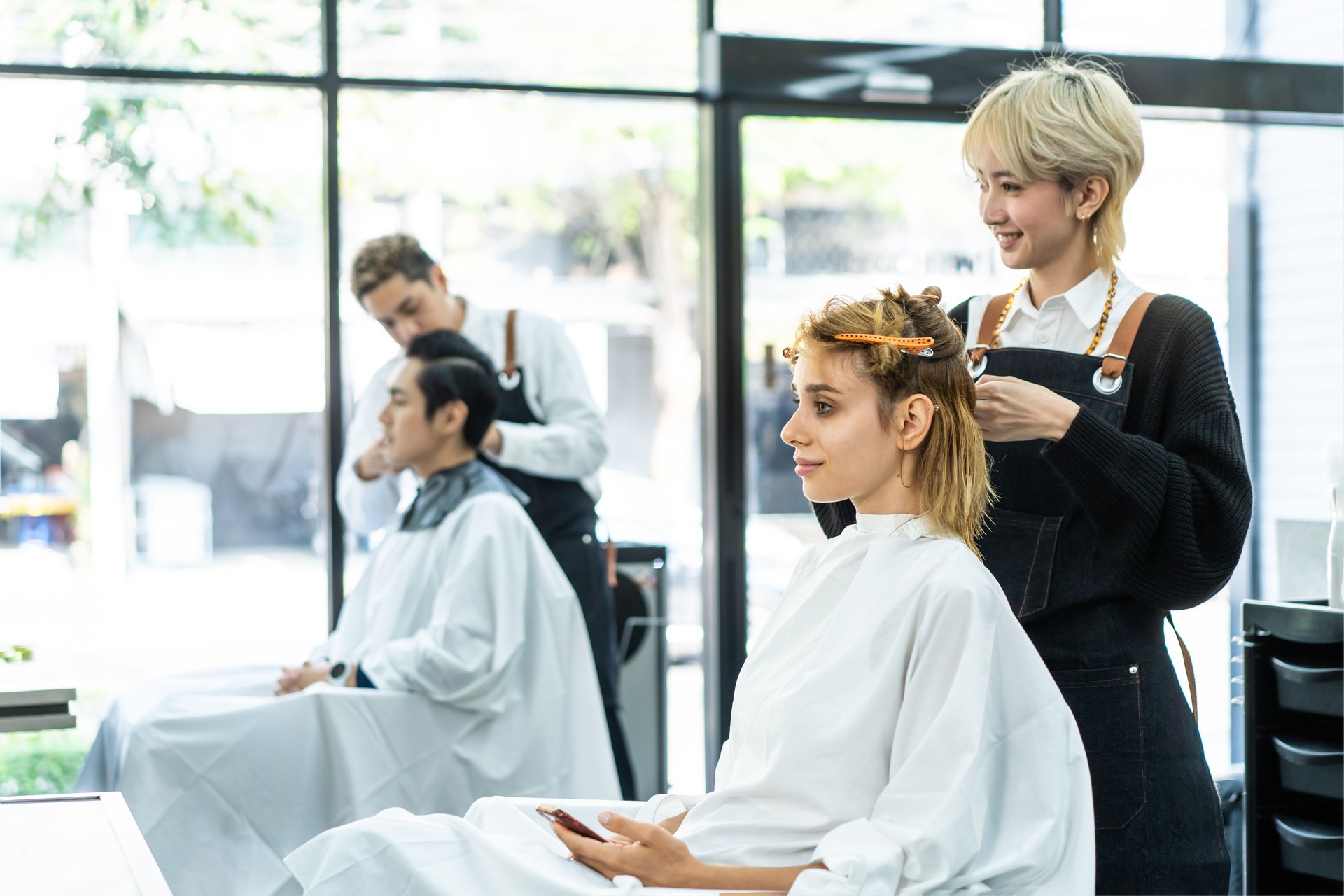 Professional young Asian female hairdresser or hair stylist talking, laughing, using scissors to cut beautiful Caucasian woman customer's blonde hair sitting with confidence in a modern beauty salon.